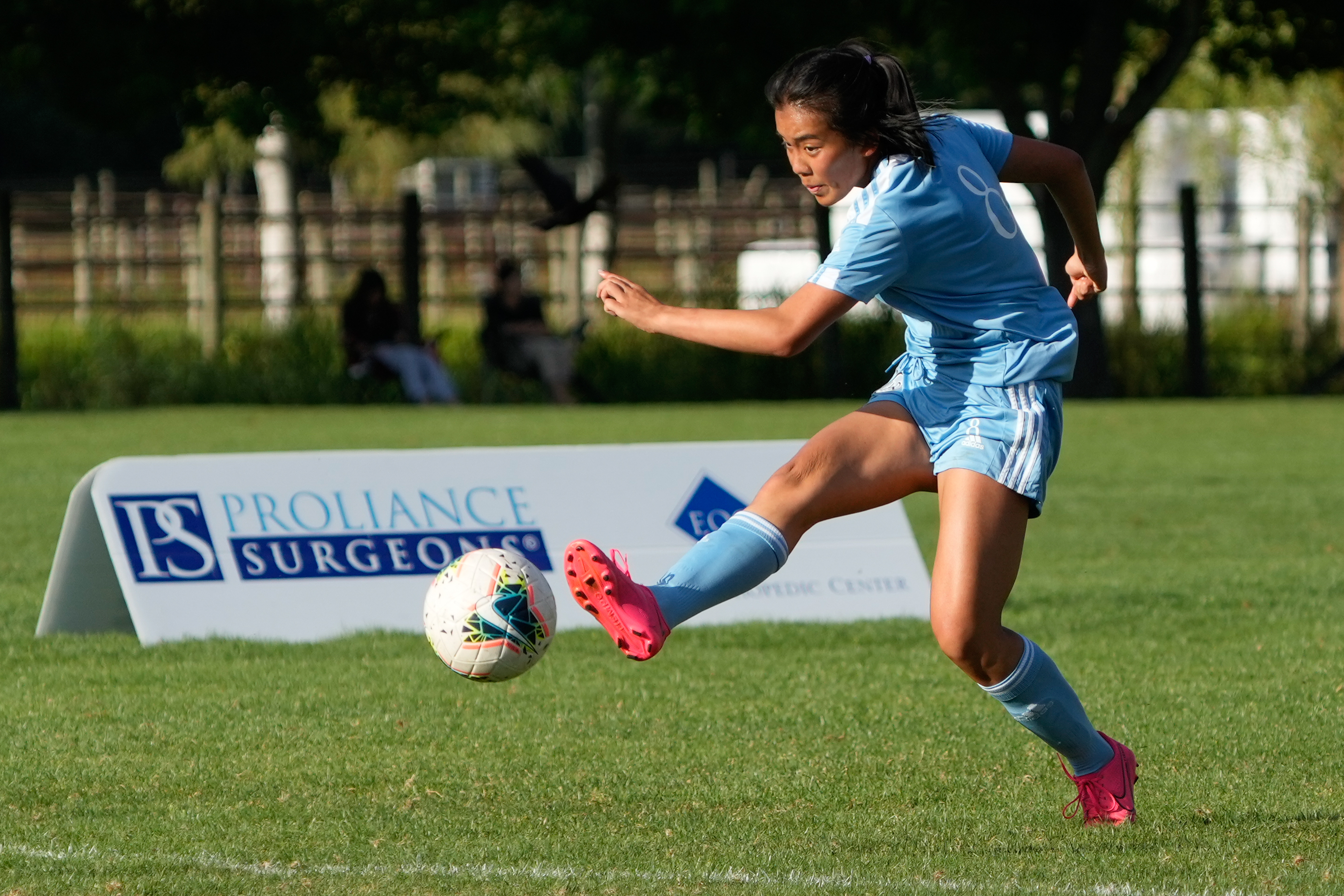 Female player wearing a light blue Adidas uniform kicking a soccer ball during a game.